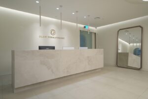 긴 대리석 리셉션 데스크 뒤 벽면에 GLOW DERMATOLOGY 로고가 붙어 있고, 천장에는 봉 형태의 펜던트 조명이 늘어져 있으며 오른쪽에는 팔각 프레임 전신 거울이 놓인 광피부과의원 청담 접수 공간입니다.