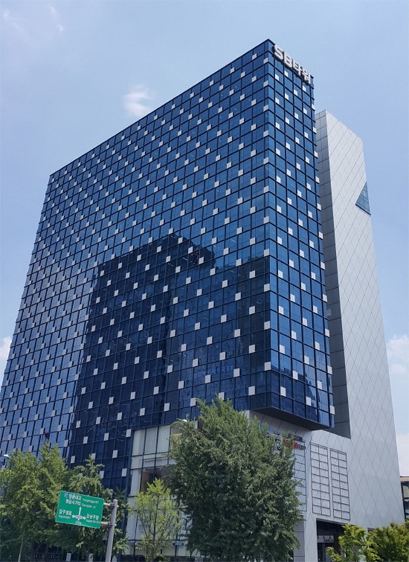 푸른 유리 외벽의 고층 건물을 아래에서 올려다본 사진으로, 앞쪽에는 가로수와 도로 안내 표지판이 함께 보이고 하늘은 맑습니다.