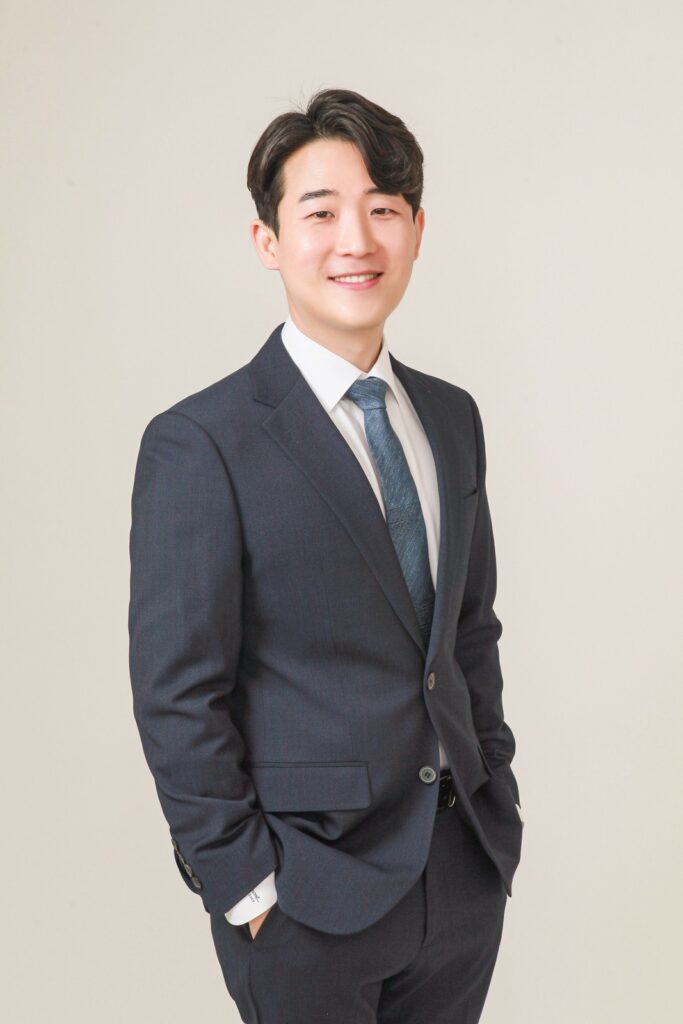 밝은 베이지색 배경 앞에서 남색 정장에 푸른색 넥타이를 맨 광피부과의원 청담 대표원장이 정면을 보고 미소 짓고 있는 프로필 사진입니다.